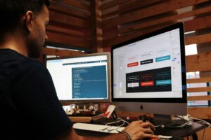 Person workin in front of a computer with 2 screens. On one screen, several colored boxes and fonts, on the other screend coding appears over a blue background. Photo by Campaign Creators on Unsplash